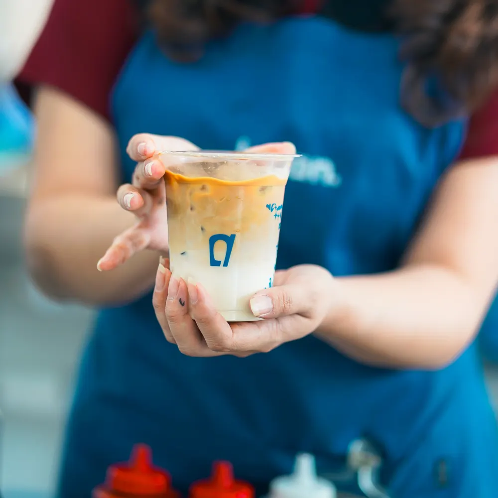 Barista Locan dengan apron biru menyodorkan es kopi susu dalam gelas Locan